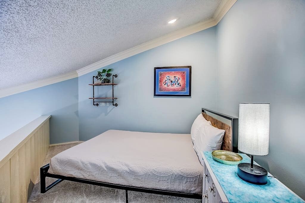 A bedroom with a bed, a bedside table with a lamp, a wall shelf with plants, and a framed picture.