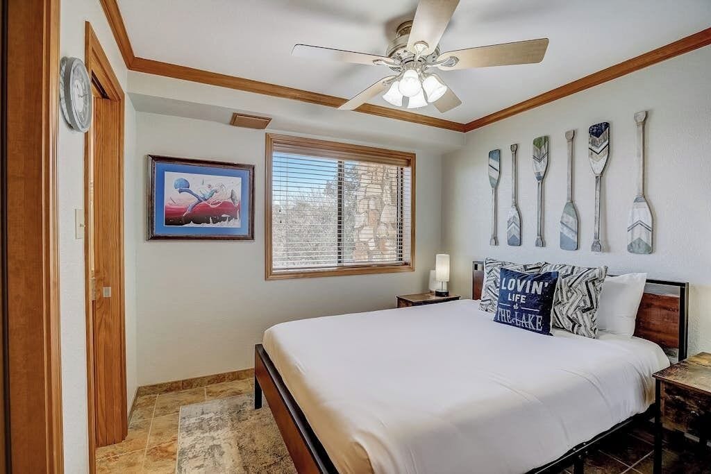 A bedroom with a bed, a ceiling fan, a window with blinds, a painting, and paddleboard decorations on the wall.