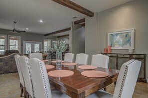 A dining room with a wooden table, white upholstered chairs, a vase with flowers, and a painting on the wall. - Hill Country Escape Hot Tub Pet Friendly (Dripping Springs)