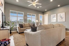 Ein Wohnzimmer mit einer Couch, einem Fernseher, einem Deckenventilator und großen Fenstern.