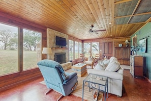 A spacious living room with a fireplace, large windows, and a ceiling fan. - Historic Lakefront & Wildlife Haven With Firepit & Gas Grill - Minutes to Water (Burnet)