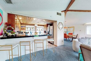 A modern kitchen with a bar area, wooden ceiling beams, and a dining area with a sofa and chairs. - Mid-century Modern Lake House With Private Waterfront Pool & Boat Dock (Marble Falls)