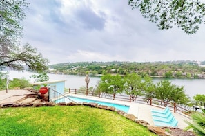 A swimming pool with a view of a lake and hills, surrounded by a stone deck and a wooden fence. - Mid-century Modern Lake House With Private Waterfront Pool & Boat Dock (Marble Falls)