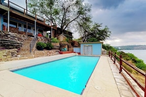 Ein Pool mit einer Steindeck, ein kleines Gebäude mit einer blauen Tür und Blick auf das Wasser.
