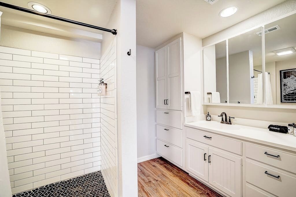A bathroom with white tiled walls, a wooden floor, a white vanity with a mirror, and a shower area with a black and white tiled floor.