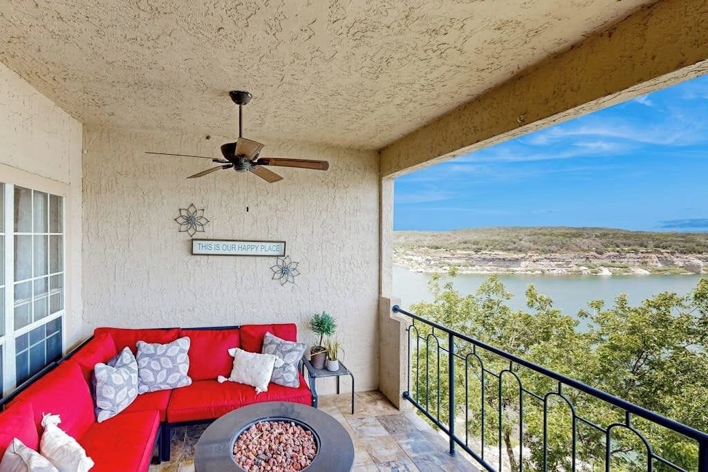A balcony with a red sofa, a ceiling fan, a fire pit, and a view of a lake.