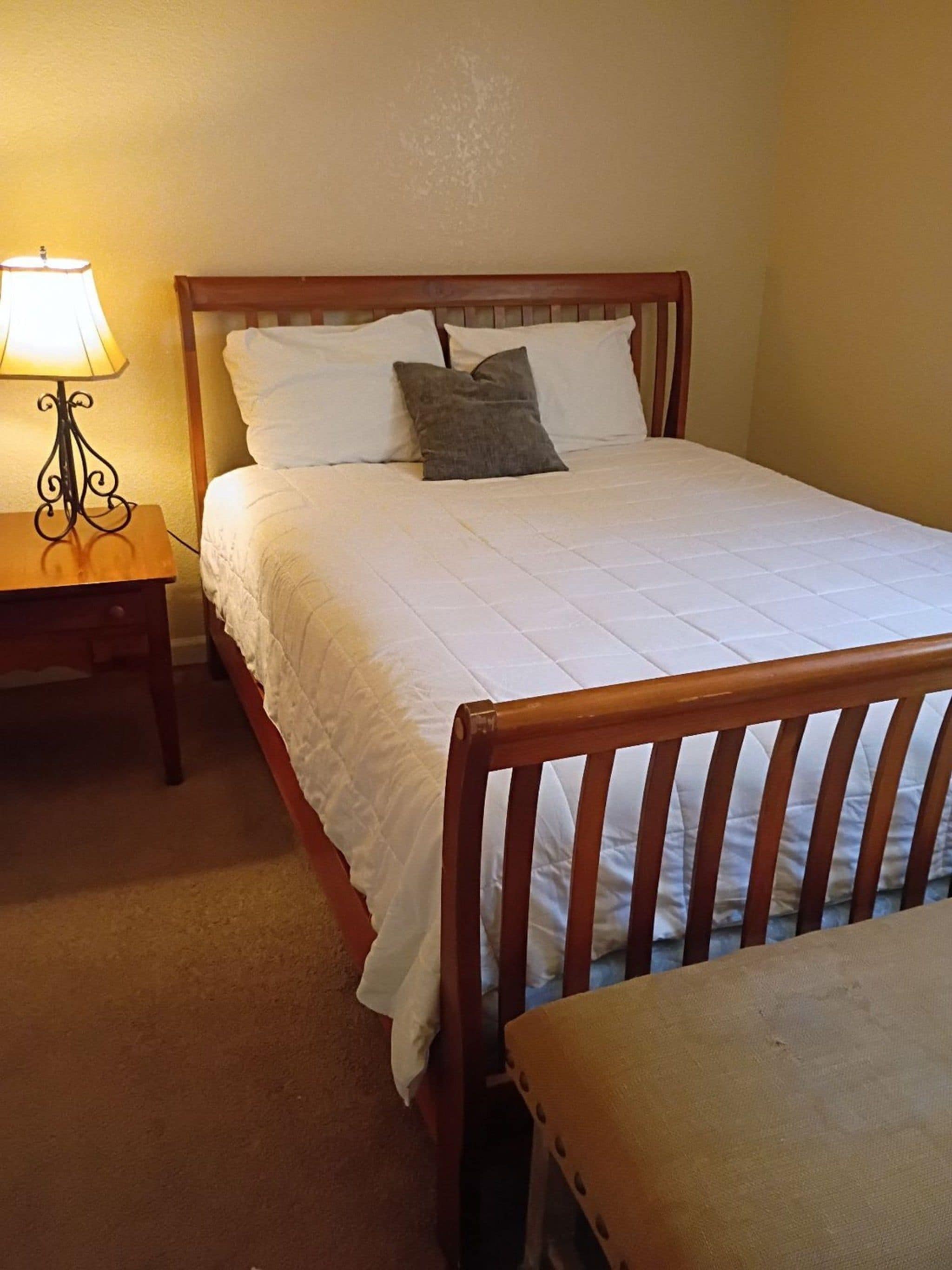 Una cama bien hecha con ropa de cama blanca y un cabecero de madera, una mesita de noche con una lámpara y una silla de madera en una habitación con paredes color beige.