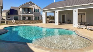 A swimming pool with a fountain feature, surrounded by a covered patio area with lounge chairs. - Stunning Lakefront Home With Pickleball, Shared Pool & Sand Volleyball (Kingsland)