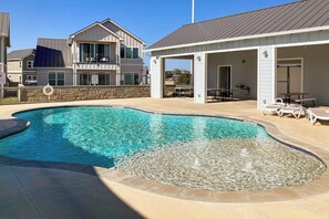 A swimming pool with a fountain feature, surrounded by a covered patio area with lounge chairs. - Stunning Lakefront Home With Pickleball, Shared Pool & Sand Volleyball (Kingsland)