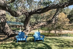 Zwei blaue Adirondack-Stühle unter einem großen Baum am Wasser.