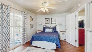 Une chambre à coucher comprenant un lit, un ventilateur de plafond, une fenêtre avec des rideaux, une table de chevet et une commode.
