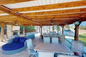 Une terrasse extérieure avec une cheminée en pierre, un canapé rond bleu, une table avec des chaises et un coin bar.