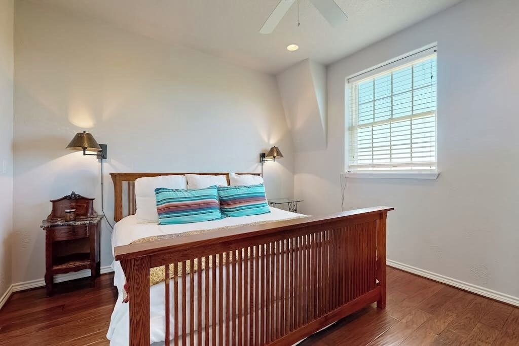 A bedroom with a wooden bed, two bedside lamps, a nightstand, a window with blinds, and a ceiling fan.
