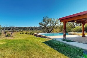 Ett poolområde med en pergola av trä, en välskött gräsmatta och utsikt över det omgivande landskapet.