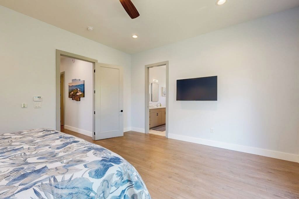 A bedroom with a bed, a ceiling fan, a flat-screen TV mounted on the wall, and a door leading to a bathroom.