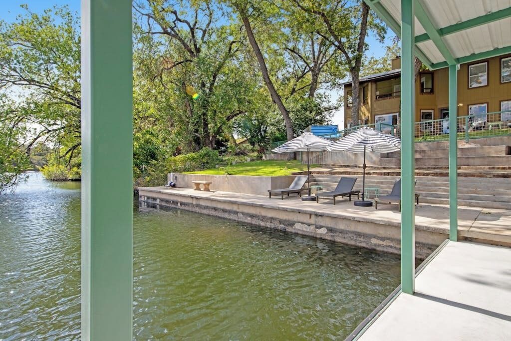 Une terrasse au bord de l’eau, dotée de chaises longues et de parasols, une piscine et un bâtiment avec des marches descendant vers l’eau.