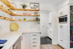 Una cocina moderna con armarios blancos, estantes de madera, un reloj y un cartel que dice 'stay AWHILE'.