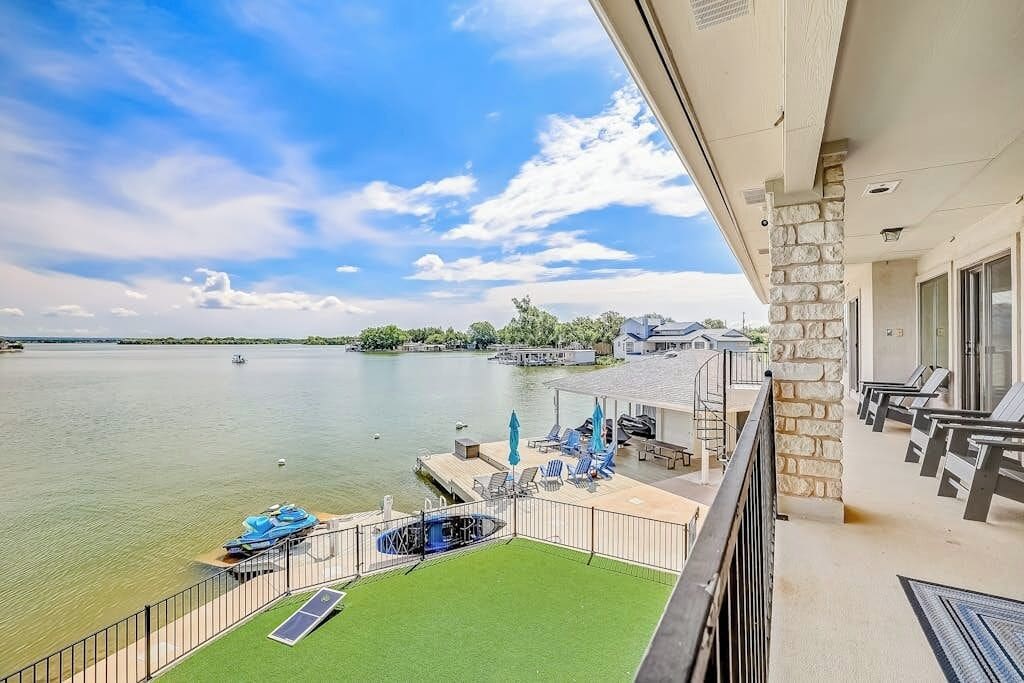 A balcony with a view of a lake, a pool, and a deck area with chairs and umbrellas.