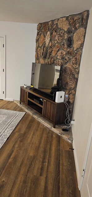 A room with a stone accent wall, a flat-screen TV on a wooden stand, a patterned rug, and a power outlet. - Central Tucson Retreat 2BR Townhome + Garage + AC for relaxing stays (Tucson)