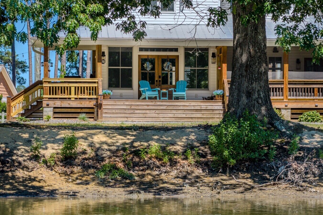 Una cabaña junto al lago con una terraza de madera, asientos exteriores y un gran árbol en primer plano.