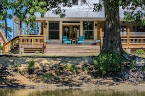 A lakeside cabin with a wooden deck, outdoor seating, and a large tree in the foreground. - Driftwood Cove Waterfront/Dock/Game house (Miles)