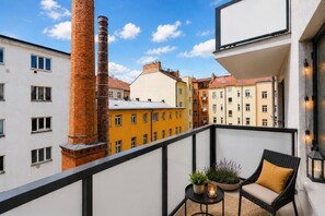 Ein Balkon mit Korbstuhl, einem kleinen Tisch mit Pflanzen und Blick auf farbenfrohe Häuser und einen Schornstein.