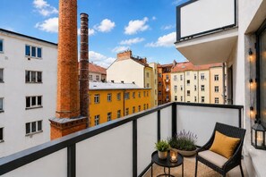 Un balcón con vistas a edificios cercanos, una silla de mimbre, una mesita con plantas y una vela.
