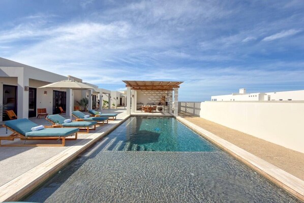 Une piscine avec des chaises longues, un espace de détente couvert et une vue sur la mer.