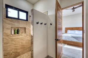 Une salle de bain moderne avec une douche, une étagère contenant des articles de toilette et une porte en bois donnant sur une chambre à coucher avec un lit.
