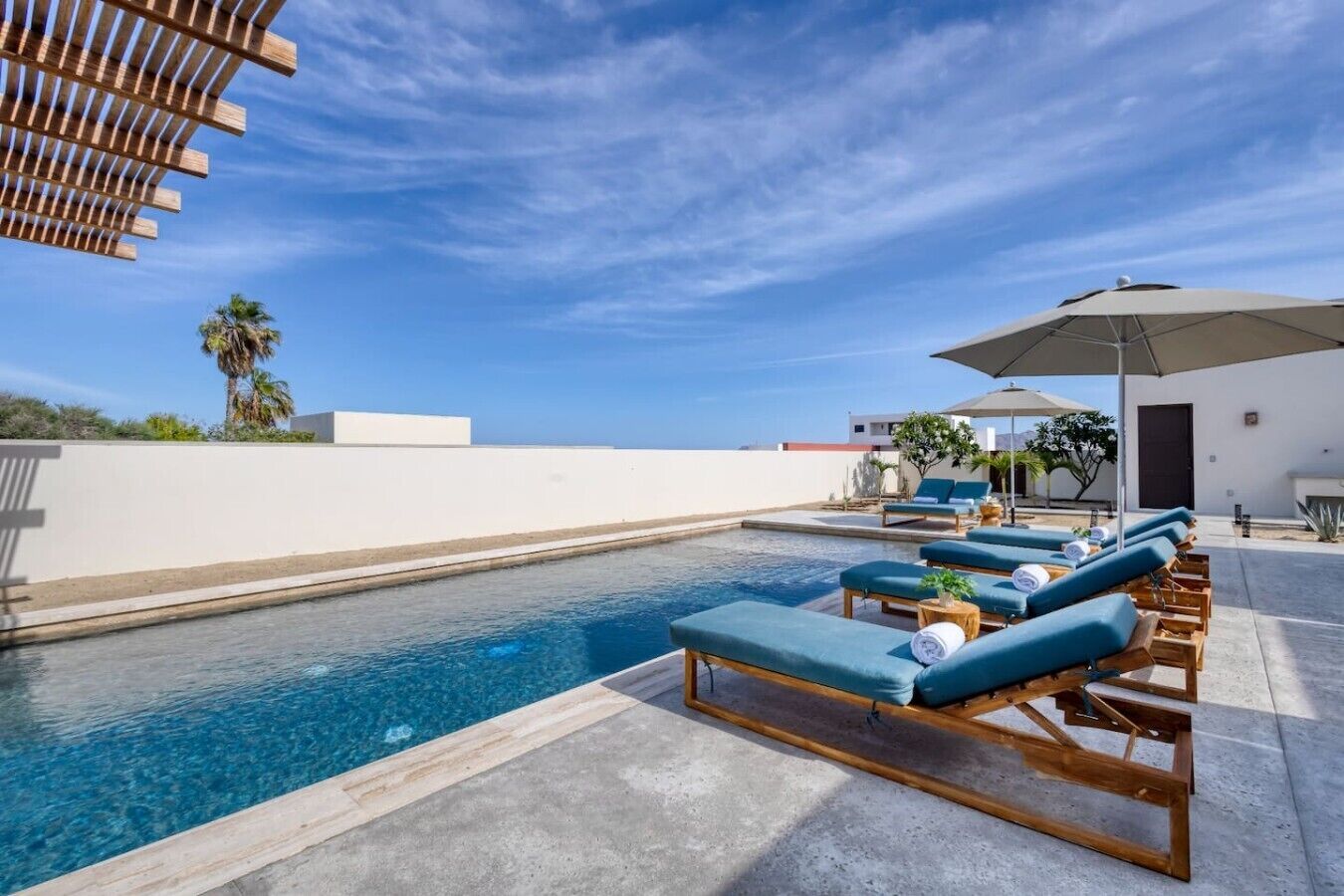 A poolside area with lounge chairs and umbrellas, a clear sky, and a white building in the background.