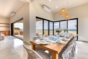A dining area with a view of the beach, a wooden table set for four, wicker chairs, and a hanging light fixture.