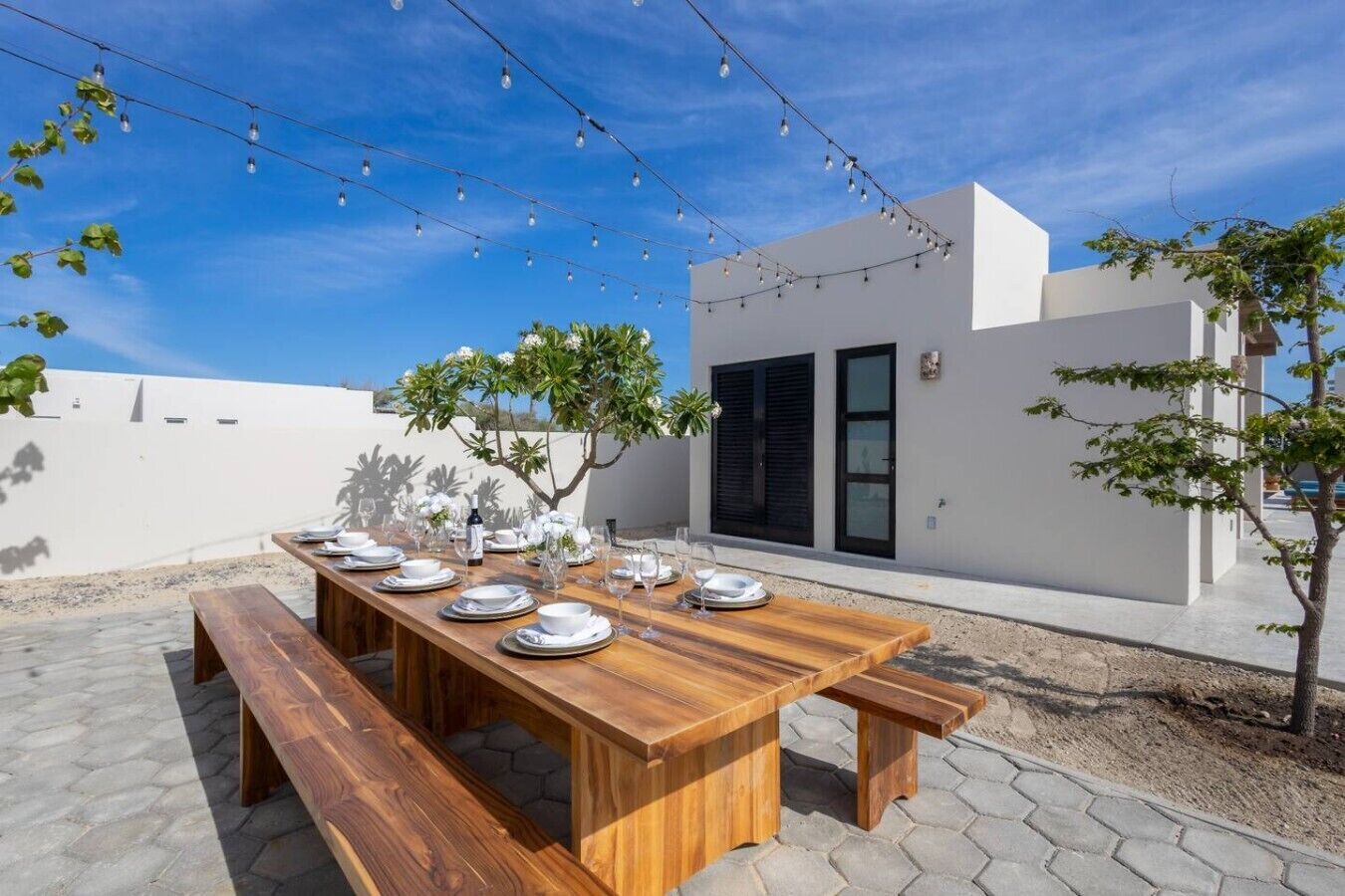 An outdoor dining area with a wooden table set for four, string lights overhead, and a modern building in the background.