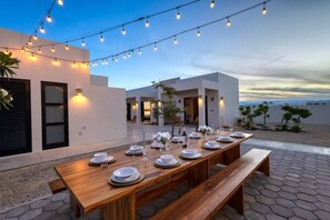 An outdoor dining area with a wooden table set for a meal, string lights overhead, and a view of the beach in the background.