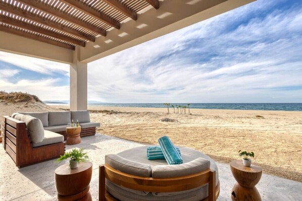 A covered outdoor seating area with a view of the beach, a sofa, a round table, and a chair.