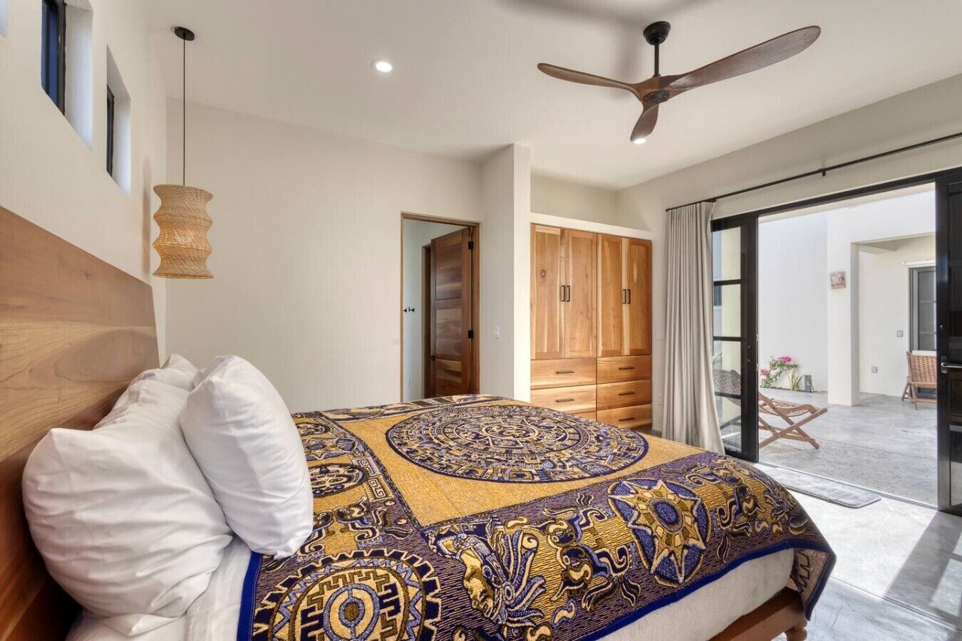 A bedroom with a large bed, a ceiling fan, a wooden wardrobe, and a view of the outside.