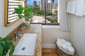 Ein Badezimmer mit Waschbecken, einem Fenster mit Blick auf die Stadt, einer Pflanze und einem Wäschekorb mit Handtüchern.