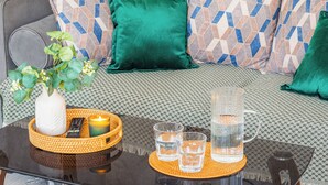 Ein Bett mit gemusterten Laken, ein grünes Kopfkissen, eine Fernbedienung, eine Kerze und Gläser auf einem Tisch.