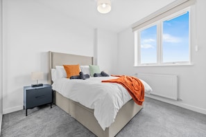 A bedroom with a bed, nightstand, lamp, and a window. - 2BR Modern Apartment | 12 Mins to Heathrow (Staines-upon-Thames)