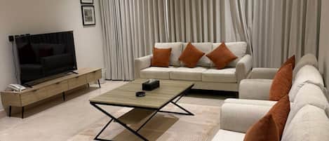 A modern living room with a sofa, a coffee table, a TV, and a large window with curtains.