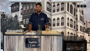 Uma área de recepção de hotel com um balcão de madeira, uma vitrine de vidro com bebidas, e um painel de fachada de edifício ao fundo.