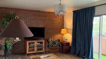 A living room with a brick wall, a television, a wooden cabinet, a patterned chair, a wooden chest, a floor lamp, and a window with curtains.