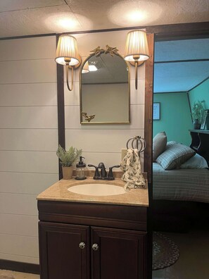 Un baño con tocador, lavamanos, dos luces fijadas a la pared, un espejo con marco decorativo y una planta en maceta.