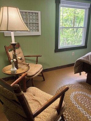 A room with a wooden chair, a small round table with a lamp, a window with a view of trees, and a bed with a patterned bedspread.
