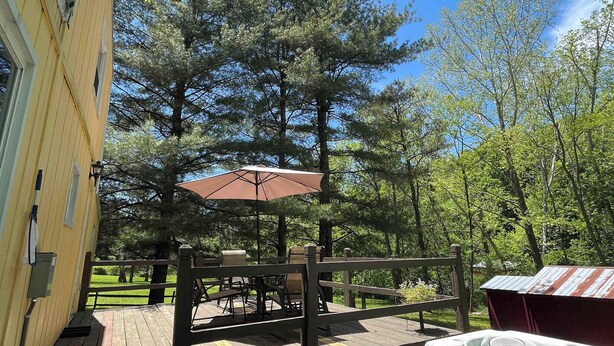 A wooden deck with a hot tub, a covered outdoor seating area, and a view of a forested area. - Charming Creekside Cottage with Hot tub (Laurelville)