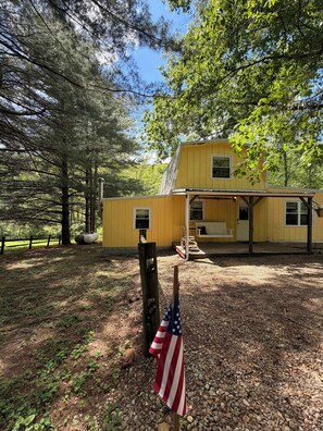 Une cabane jaune avec un porche, entourée d’arbres et bordée d’un sol en gravier.