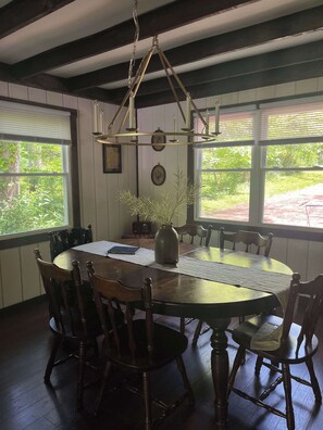 Une salle à manger avec une table ronde, des chaises en bois, un lustre et de grandes fenêtres équipées de stores.