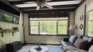 A cozy living room with a ceiling fan, a round wooden coffee table, a sofa, and a large window with a view of greenery. - Charming Creekside Cottage with Hot tub (Laurelville)