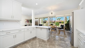 A modern kitchen with white cabinetry, a large island, and a dining area with a view of the outdoors. - Premier 3-Bedroom Condo in Coveted Indian Wells Country Club, Discounted Rates! (Indian Wells)