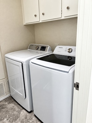 A laundry room with two white front-loading washers and dryers, a beige wall, and a white door. - Premier 3-Bedroom Condo in Coveted Indian Wells Country Club, Discounted Rates! (Indian Wells)