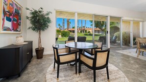 A dining area with a round table, four chairs, a potted plant, a cabinet, and a large window with a view of palm trees and a lawn. - Premier 3-Bedroom Condo in Coveted Indian Wells Country Club, Discounted Rates! (Indian Wells)